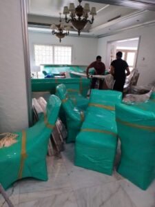 لقطة لغرفة داخلية أثناء عملية الانتقال، تظهر فيها كراسي وقطع أثاث مغلفة بالكامل بغطاء بلاستيكي واقي باللون الفيروزي (اللبني)، وفي الخلفية يظهر شخصان يقومان بنقل الأثاث.
